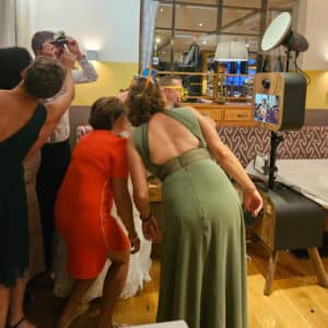 Eine Gruppe von Menschen in farbenfroher, formeller Kleidung steht dicht beieinander, wirft sich in verspielte Posen und trägt lustige Brillen, während sie ein Foto mit einem Fotoautomaten bei einer Indoor-Veranstaltung machen.