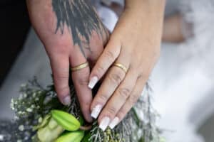 Zwei Hände mit goldenen Eheringen ruhen auf einem Blumenstrauß; eine Hand hat eine schwarze Tätowierung und die andere eine weiße Nagelmalerei mit einem kleinen Muster. Unter den Händen sind grüne Blumen und weißes Laub zu sehen.