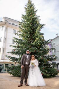 Braut und Bräutigam in Hochzeitskleidung stehen lächelnd vor einem großen geschmückten Weihnachtsbaum in einer städtischen Umgebung im Freien, mit Gebäuden im Hintergrund.