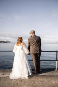 Eine Braut im weißen Kleid und ein Bräutigam im braunen Anzug halten sich an der Hand, stehen an einem Metallgeländer und schauen sich an einem ruhigen Gewässer unter einem klaren Himmel an.