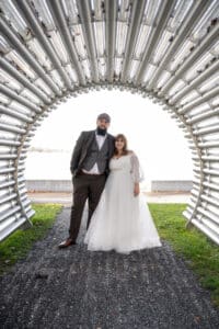 Eine Braut in einem weißen Kleid und ein Bräutigam im Anzug stehen lächelnd zusammen unter einem Tunnel aus gebogenen Metallrohren, im Hintergrund sind grünes Gras und ein See zu sehen.