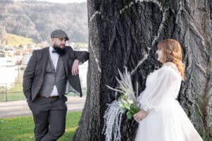 Ein Bräutigam in einem Anzug und einer flachen Kappe lehnt an einem großen Baum und lächelt eine Braut in einem weißen Kleid an, die einen Blumenstrauß hält. Sie stehen im Freien mit Hügeln und Wasser im Hintergrund.