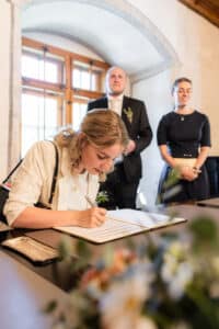 Eine Frau mit blondem Haar unterschreibt ein Dokument an einem Tisch, während im Hintergrund in einem hellen, sonnenbeschienenen Raum zwei Personen stehen und lächeln. Die Blumen im Vordergrund sind unscharf.