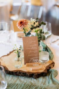 Ein Hochzeitstisch mit einem hölzernen Tafelaufsatz, Glasvasen mit Blumen, einem gedruckten Menü, Kerzen und einem klaren Acrylschild mit der Aufschrift "Tisch sieben" in eleganter Schrift.