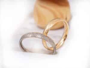 Zwei kunstvoll gravierte Ringe, ein silberner und ein goldener, stehen nebeneinander auf einer weißen Fläche mit einem verschwommenen, hellbraunen Objekt im Hintergrund.