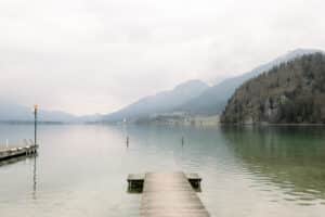 Ein hölzerner Steg ragt in einen ruhigen, klaren See, umgeben von nebligen Bergen und bewaldeten Hügeln unter einem wolkenverhangenen Himmel. Das Wasser ist ruhig, und das ferne Ufer ist nur schwach zu erkennen.