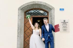 Eine fröhliche Braut in einem weißen Hochzeitskleid und ein Bräutigam in einem blauen Anzug halten sich vor einem Gebäude an den Händen. Die Braut hebt ihren Brautstrauß und lächelt, während der Bräutigam eine rote Mappe hält. Sie stehen vor einer Holztür und einem Schild.