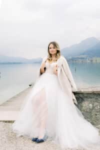 Eine Frau in einem weißen Hochzeitskleid und einem hellen Mantel sitzt auf einem Steinsockel an einem See, mit Bergen und einem bewölkten Himmel im Hintergrund. Sie trägt blaue Schuhe und schaut ruhig in die Kamera.