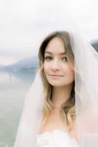 Eine junge Braut mit gewelltem blondem Haar und blauen Augen, die einen weißen Schleier trägt, steht im Freien in der Nähe eines ruhigen Gewässers mit Bergen im Hintergrund. Sie hat ein sanftes Lächeln und trägt ein weißes Hochzeitskleid.