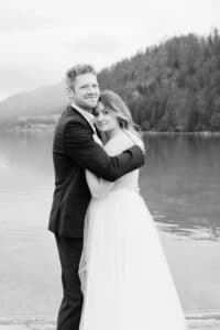 Eine Braut und ein Bräutigam umarmen sich auf einem Steg an einem ruhigen See, umgeben von Bergen. Die Braut trägt ein fließendes weißes Kleid und der Bräutigam einen dunklen Anzug. Das Foto ist in schwarz-weiß gehalten.
