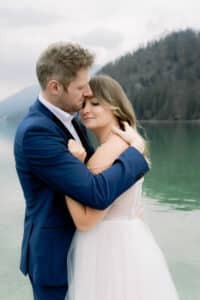 Ein Mann in einem marineblauen Anzug umarmt und küsst eine Frau in einem weißen Kleid an einem ruhigen See, mit baumbewachsenen Bergen im Hintergrund. Die Frau hat die Augen geschlossen und lehnt sich in den zärtlichen Moment hinein.