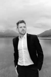 Ein Mann im Anzug steht an einem ruhigen See mit Bergen im Hintergrund unter einem bewölkten Himmel und schaut nachdenklich in die Ferne. Das Bild ist in schwarz-weiß gehalten.