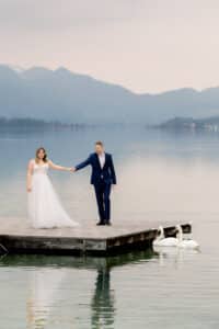 Eine Braut in einem weißen Kleid und ein Bräutigam in einem blauen Anzug stehen Hand in Hand auf einem hölzernen Steg über einem ruhigen See, mit Bergen im Hintergrund und zwei Schwänen, die in der Nähe schwimmen.