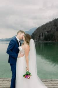 Eine Braut und ein Bräutigam stehen auf einem hölzernen Steg an einem ruhigen See und teilen einen intimen Moment. Die Braut hält einen Blumenstrauß und trägt ein weißes Kleid mit Schleier, während der Bräutigam einen marineblauen Anzug trägt. Im Hintergrund sind bewaldete Hügel und ein bewölkter Himmel zu sehen.