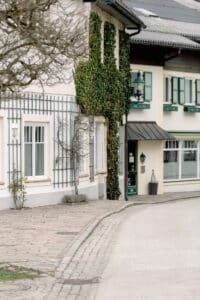 Eine malerische europäische Straße mit hellen Gebäuden, grünen Fensterläden und Efeu an einer Fassade. Ein kahler Baum steht auf dem Gehweg, und die Szene wirkt ruhig und still.