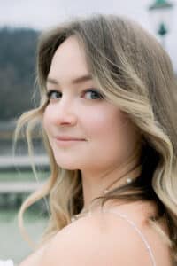 Eine junge Frau mit gewelltem blondem Haar und heller Haut lächelt sanft in die Kamera im Freien. Sie trägt ein zartes weißes Kleid und eine Perlenkette. Im verschwommenen Hintergrund sind Grünpflanzen und eine Laterne zu sehen.