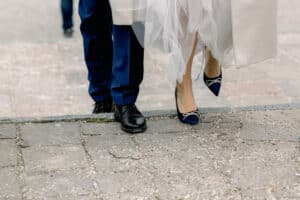 Nahaufnahme eines Paares, das im Freien spazieren geht. Der Mann trägt eine dunkelblaue Hose und schwarze Schuhe, die Frau ein weißes Kleid mit marineblauen Absätzen. Beide treten auf einen Steinbordstein.