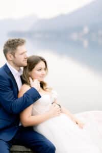 Ein Paar sitzt eng beieinander an einem ruhigen See, mit Bergen und einem dunstigen Himmel im Hintergrund. Der Mann trägt einen marineblauen Anzug und die Frau ein weißes Kleid. Beide wirken entspannt und zufrieden und genießen die friedliche Landschaft.