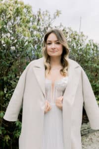 Eine Frau in einem weißen, trägerlosen Kleid und einem hellbeigen Mantel steht im Freien vor grünen, belaubten Sträuchern und blickt mit sanftem Ausdruck in die Kamera.