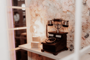Ein altes Telefon mit Drehscheibe steht auf einem weißen Regal neben einer leuchtenden Glühbirne und einer kleinen Holzkiste, mit einer blumengemusterten Wand im Hintergrund.