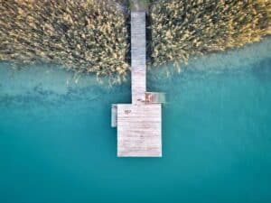 Luftaufnahme eines hölzernen Stegs, der sich über ruhiges, türkisfarbenes Wasser erstreckt und von hohem Schilf oder Gräsern am Ufer gesäumt wird. Der Steg bildet eine T-Form, deren Schatten im klaren Wasser sichtbar sind.