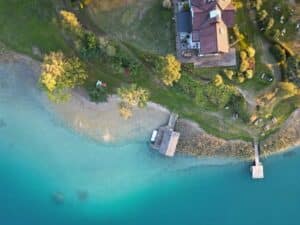 Luftaufnahme eines Hauses mit rotem Dach an einem türkisfarbenen See, umgeben von grünen Bäumen und Gras, mit einem kleinen Steg und einem Bootshaus, das sich über das Wasser erstreckt.