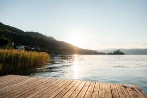 Ein hölzerner Steg überblickt einen ruhigen See bei Sonnenuntergang, mit Schilf auf der linken Seite, entfernten Bergen und einem kleinen Dorf am Seeufer unter einem klaren Himmel.