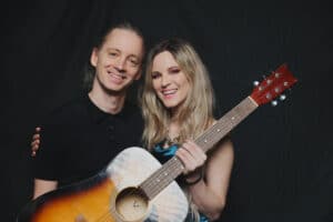 Ein lächelnder Mann in einem schwarzen Hemd und eine lächelnde Frau mit langen blonden Haaren, die eine Akustikgitarre in der Hand hält, stehen zusammen vor einem dunklen Hintergrund.