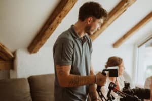 Ein Mann mit Tätowierungen an den Armen bedient eine professionelle Kameraausrüstung in einem Innenraum und steht an einem Fenster mit freiliegenden Holzbalken an der Decke. Eine Frau sitzt im Hintergrund auf einer Couch.