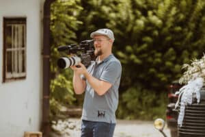 Ein Mann mit einem hellblauen Hemd, einer Mütze und einer Brille filmt im Freien mit einer professionellen Videokamera. Im Hintergrund sind Grünflächen und ein Teil eines dekorierten Fahrzeugs zu sehen.