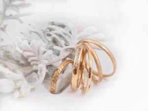 Vier goldene Ringe, jeder mit einem einzigartigen Design, sind auf einem weichen, weißen, gefrosteten Hintergrund mit gefiedertem Laub und einer verträumten, eleganten Atmosphäre angeordnet.