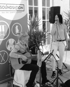 Eine Frau singt in ein Mikrofon, während ein Mann neben ihr eine akustische Gitarre spielt. Sie treten im Freien neben einem "SOUNDSATION"-Musikbanner auf, mit Topfpflanzen und Sonnenlicht im Hintergrund.