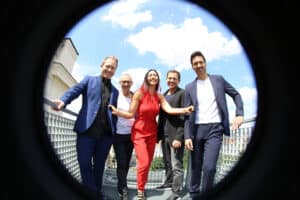 Fünf halbförmig gekleidete Personen lächeln und posieren zusammen auf einem Balkon im Freien, wobei ein Fischaugenobjektiv das Bild vor einem hellen, teilweise bewölkten Himmel einrahmt.