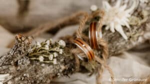 Zwei hölzerne Ringe mit einem kleinen Diamanten sind mit Schnur an einem rustikalen, mit weißen Blumen und Grünzeug geschmückten Zweig zusammengebunden. Der Hintergrund ist sanft verschwommen. Bildnachweis: Jennifer Grünauer.