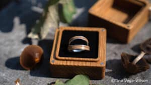 Ein Paar Ringe aus Silber und Holz ruht in einer kleinen hölzernen Ringschachtel, umgeben von getrockneten Blättern und Pilzen auf einer sonnenbeschienenen Holzfläche. ©Tina Vega-Wilson.
