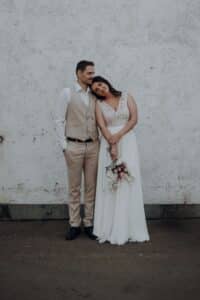 Eine Braut in einem weißen Kleid lehnt ihren Kopf an die Schulter des Bräutigams, während sie gemeinsam vor einer strukturierten weißen Wand stehen. Sie hält einen Blumenstrauß in der Hand, und die beiden sehen friedlich und zufrieden aus.