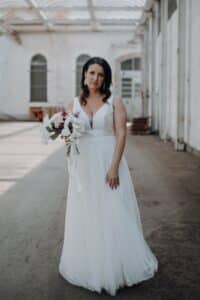 Eine Frau in einem weißen Hochzeitskleid mit einem Blumenstrauß in der Hand steht in einem sonnendurchfluteten Flur im Industriestil mit hohen Fenstern und einer Glasdecke. Sie blickt ruhig in die Kamera.
