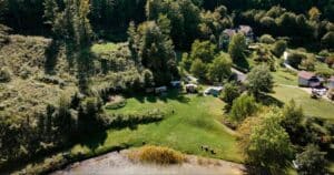 Luftaufnahme einer grasbewachsenen Lichtung mit mehreren kleinen Gebäuden, Zelten und Fahrzeugen, umgeben von dichtem Wald und einem Teich im Vordergrund. Zwischen den Bäumen im Hintergrund sind einige Häuser zu sehen.