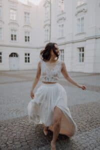 Eine Frau in einem ärmellosen weißen Spitzenoberteil und einem wallenden Rock läuft fröhlich über einen Hof mit Kopfsteinpflaster und weißen historischen Gebäuden im Hintergrund.