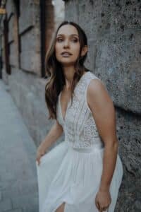 Eine Frau in einem ärmellosen weißen Spitzenkleid mit fließendem Rock steht an einer Steinmauer und blickt mit ruhiger Miene leicht nach oben. Ihr langes braunes Haar fällt ihr über die Schulter.