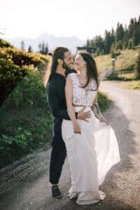 Ein Paar steht auf einem sonnenbeschienenen Weg in der Natur; der Mann in dunkler Kleidung umarmt die lächelnde Frau, die ein weißes Kleid trägt. Im Hintergrund sind Bäume und Berge zu sehen.