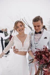 Eine lächelnde Braut in einem weißen, langärmeligen Spitzenoberteil und fließendem Rock steht neben einem Bräutigam in einem hellen karierten Anzug, der einen Strauß roter Blumen hält. Im Hintergrund sind Schnee und mehrere Personen zu sehen.