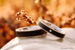 Zwei Eheringe mit einem dunklen Außenring und einem einzelnen kleinen Diamanten auf dem einen, die zusammen auf einer glatten, hellen Oberfläche mit einem verschwommenen, goldbraunen natürlichen Hintergrund liegen.