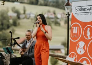 Eine Frau in einem leuchtend orangefarbenen Overall singt im Freien in ein Mikrofon, während ein Mann neben ihr Gitarre spielt. Hinter ihnen ist ein Schild mit Musiksymbolen und dem Wort "SOUNDSATION" zu sehen. Im Hintergrund ist eine malerische, grüne Landschaft zu sehen.