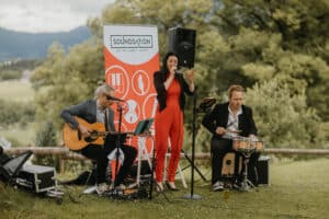 Eine Frau singt in ein Mikrofon, während zwei Männer im Freien neben einem "Soundstation"-Banner Gitarre und Schlagzeug spielen, mit Bäumen und Bergen im Hintergrund.