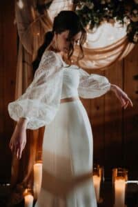 Eine Frau in einem weißen, langärmeligen Spitzenoberteil und einem dazu passenden Rock posiert anmutig in einem Innenraum, umgeben von brennenden Stumpenkerzen und drapierten Stoffen, wobei sie von einem weichen, warmen Licht angestrahlt wird.