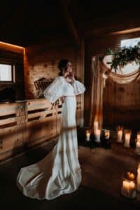 Eine Braut in einem langen weißen Kleid mit durchsichtigen Puffärmeln steht nachdenklich in einem rustikalen Holzzimmer, umgeben von brennenden Kerzen und sanftem Tageslicht, mit Grünzeug und Stoffdekorationen im Hintergrund.