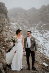 Eine Braut in einem wallenden weißen Kleid und ein Bräutigam in einer grauen Jacke gehen Hand in Hand auf einem felsigen, schneebedeckten Bergpfad und lächeln sich an.