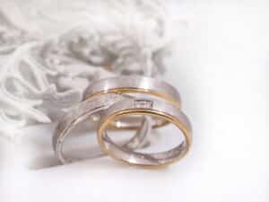 Zwei elegante Eheringe in Silber und Gold ruhen auf einer weichen weißen Oberfläche. In einem Ring ist eine kleine Reihe von Diamanten in den Ring eingefasst. Zartes weißes Blattwerk schmückt den Hintergrund.