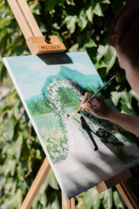 Eine Person malt eine Braut und einen Bräutigam auf einer Leinwand, die auf einer Staffelei im Freien steht, umgeben von Grün. Das Gemälde zeigt eine Hochzeitsszene mit Bergen, Blumen und einem Blumenbogen im Hintergrund.